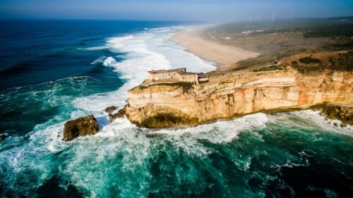 aerial image of the cliff and lighthouse in nazaré portugal. blue water crushes the rocks